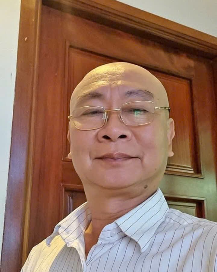 Anh Phan Khánh mặc áo sơ mi trắng sọc, đứng trước cánh cửa gỗ